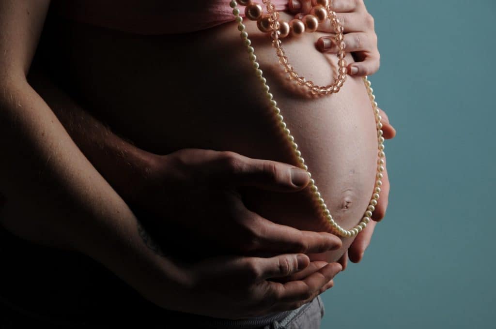 Nackter hochschwangerer Bauch wird von hinten umfasst von den Händen der Mutter und denen eines Mannes. Über dem Bauch hängen drei Perlenketten.