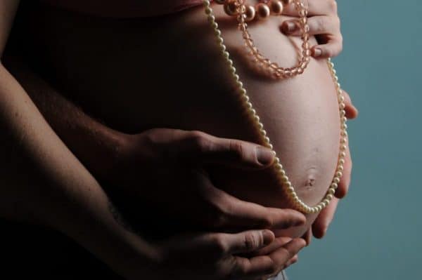 Nackter hochschwangerer Bauch wird von hinten umfasst von den Händen der Mutter und denen eines Mannes. Über dem Bauch hängen drei Perlenketten.