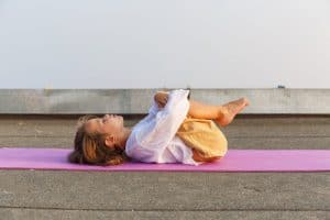 Kinder Yoga. Kind liegt auf einer Yogamatte mit angezogenen Beinen auf dem Rücken und entspannt.