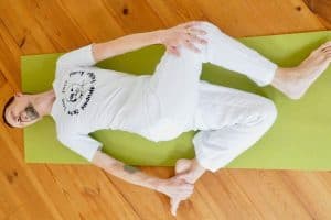 Mann, gekleidet in weiß, liegt auf einer grünen Yogamatte in einer seitlichen Drehung